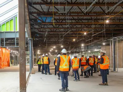 Group of people in orange safety vests and hard hats standing inside a large, unfinished construction site with exposed steel beams and concrete floors.